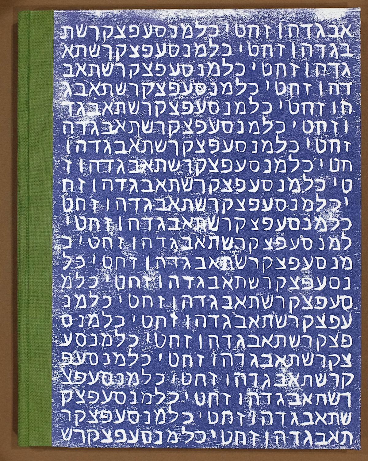 The rear cover of a handmade composition style notebook fills most of the frame. Since the notebook opens right to left, the spine is on the left side, covered in green bookcloth. The cover is a handmade relief print with a dark blue background and the Hebrew alphabet in white on repeating lines. The alphabet is written with each line offset from the previous, so Alef is first on the first line, Bet is first on the second line, and so on.