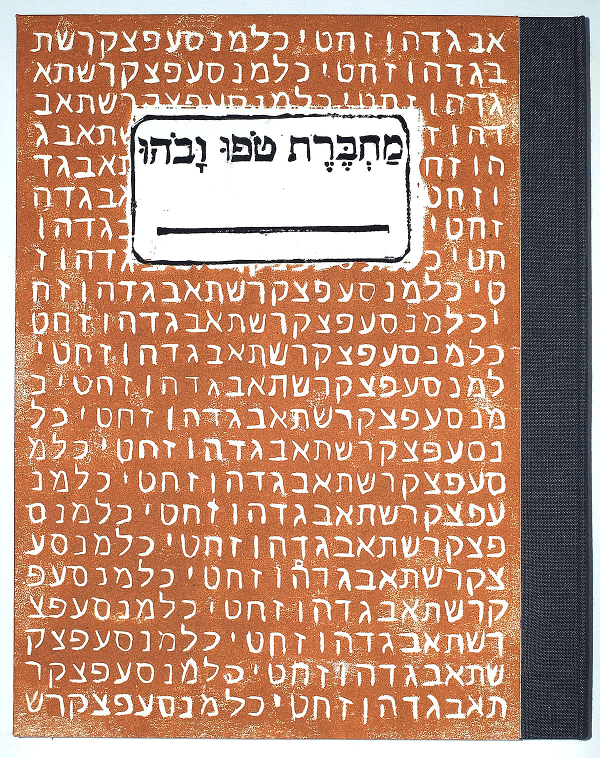 A handmade composition style notebook is shown with a grey bookcloth spine and a handmade relief print of the Hebrew alphabet with white letters on a yellow-orange-brown background. The center of the print is a white block with a black framing box, a line for writing, and black text that says "Machberet Tofu Vavohu" in Hebrew letters.