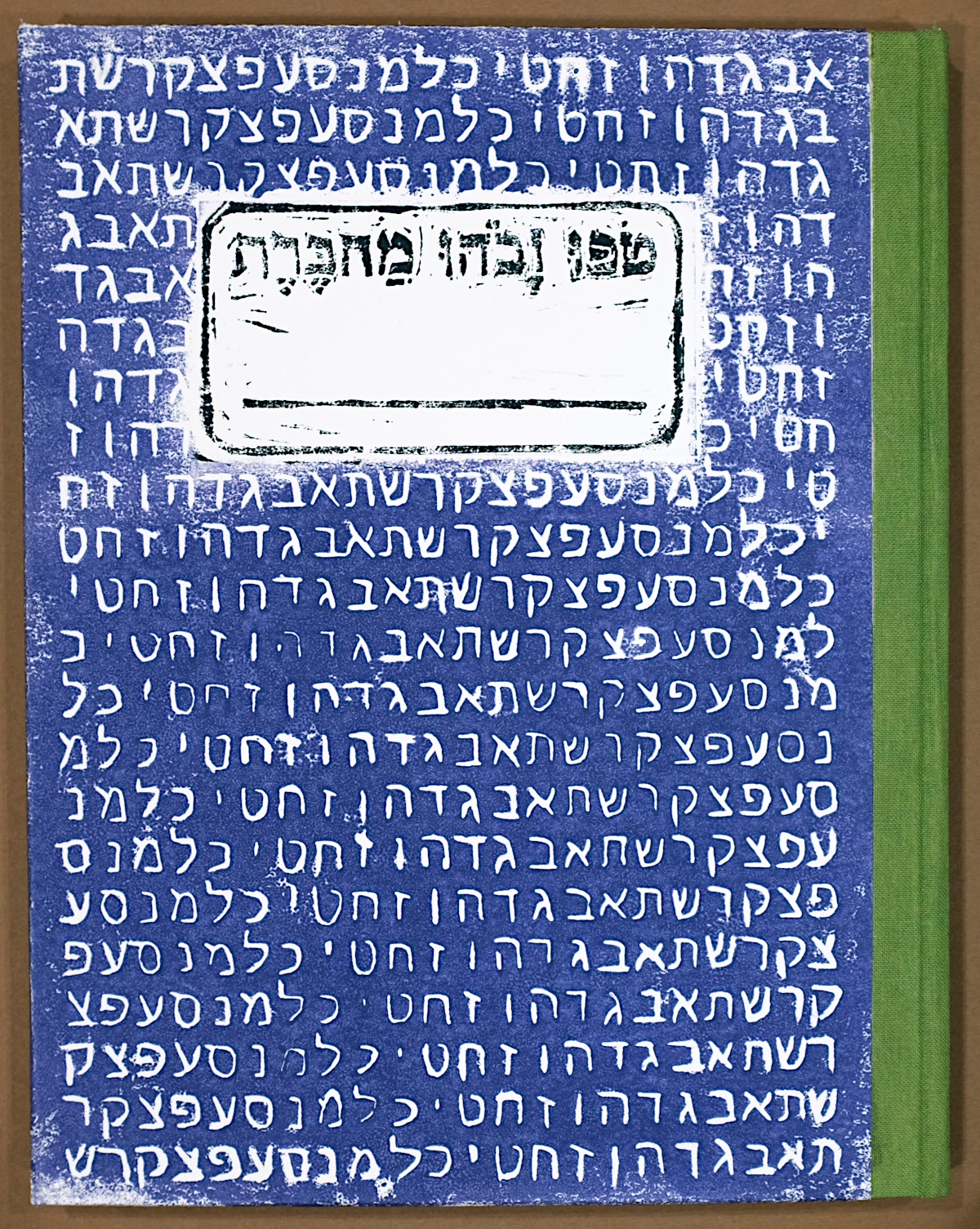 The front cover of a handmade composition style notebook fills most of the frame. Since the notebook opens right to left, the spine is on the right side, covered in green bookcloth. The cover is a handmade relief print. The center of the print is a white block with a black framing box, a line for writing, and black text that says "Tofu Vavohu Machberet" in Hebrew letters. Most of the print is a medium blue background with the Hebrew alphabet in white on repeating lines. The alphabet is written with each line offset from the previous, so Alef is first on the first line, Bet is first on the second line, and so on.